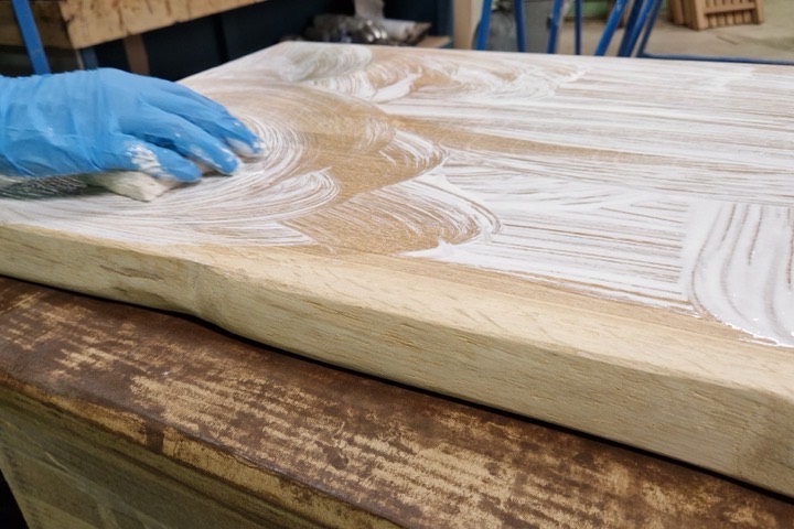 Tka home world maatwerk - Teakea Een hand met handschoen brengt afwerking aan op een houten blad in de meubelmakerij